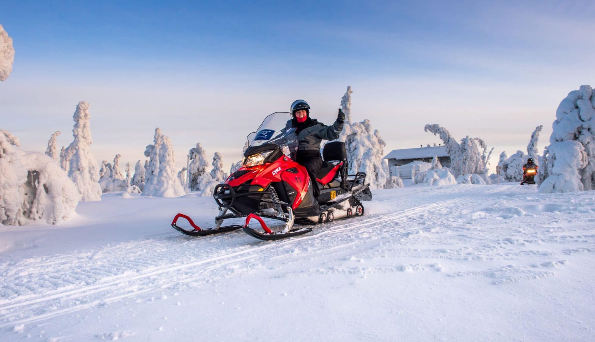 Moottorikelkkailija ajamassa lumisessa maisemassa kirkkaana talvipäivänä, taustalla lumipeitteisiä puita