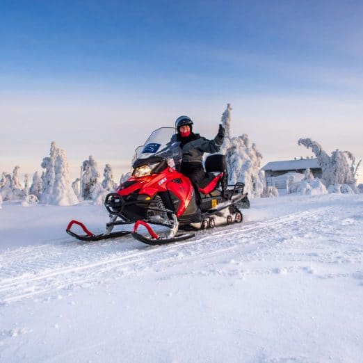 Moottorikelkkailija ajamassa lumisessa maisemassa kirkkaana talvipäivänä, taustalla lumipeitteisiä puita